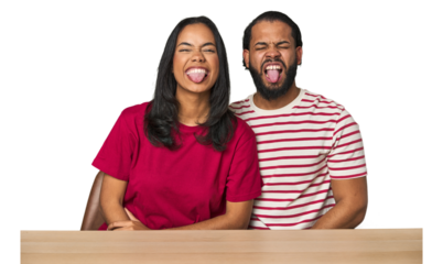 Seated young Latino couple at table funny and friendly sticking out tongue.