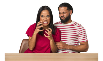 Seated young Latino couple at table upset screaming with tense hands.
