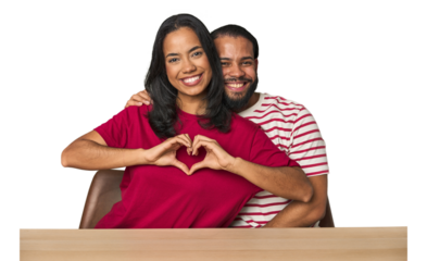 Seated young Latino couple at table smiling and showing a heart shape with hands.