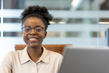 Obraz premium Young African American businesswoman smiling confidently while sitting at workplace inside office. Modern workspace with professional company employee. Person working with computer and phone