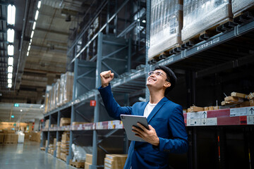 A man in a blue suit is holding a tablet and smiling. He is in a warehouse and he is happy