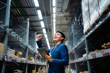 A man in a blue suit is holding a tablet and smiling. He is in a warehouse and he is happy