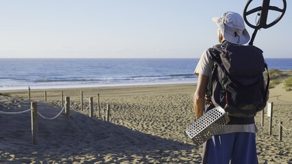 Young adult walking on a marked path towards the beach, carrying a metal detector on his shoulder...