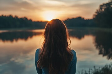 Fototapeta premium A woman stands alone by the shore of a serene lake as the sun sets behind her