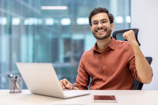 Smiling young professional celebrating success in modern office with laptop and phone on desk. Man expressing joy and achievement in business environment.