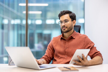 Serious businessman working with laptop and tablet in modern office. Focused on tasks, using both devices to enhance productivity. Professional setting with large windows and natural light.