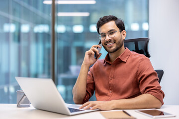 Smiling businessman in glasses talking on phone while working at desk with laptop in modern office....
