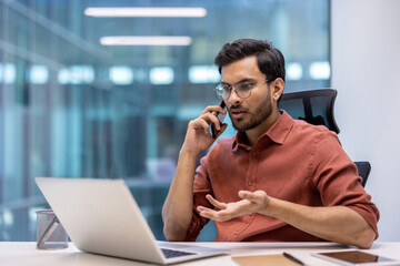 Businessman talking on phone while working on laptop in modern office setting. Professional engaged in conversation, multitasking in workspace. Concept of communication, technology, and business.