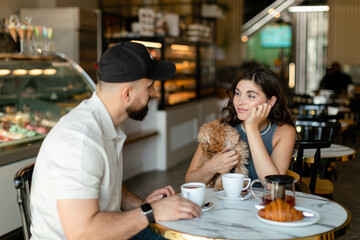 A woman with long hair with brown maltipoo and a bearded man in a cap are talking and eating croissants and drinking tea in cafe. Couple in love with little dog having breakfast in cafe.