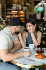 A woman with long hair with brown maltipoo and a bearded man in a cap are talking and eating croissants and drinking tea in cafe. Couple in love with little dog having breakfast in cafe.