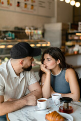A woman with long hair with brown maltipoo and a bearded man in a cap are talking and eating croissants and drinking tea in cafe. Couple in love with little dog having breakfast in cafe.