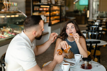 A woman with long hair with brown maltipoo and a bearded man in a cap are talking and eating croissants and drinking tea in cafe. Woman with long hair is eating croissant in cafe with her boyfriend.  
