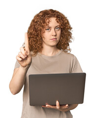 Young Caucasian redhead woman with laptop showing number one with finger.