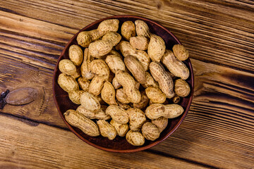 Ceramic plate with heap of unpeeled peanuts on wooden table. Top view