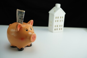 Horizontal detail photo of a piggy bank with several yen bills inserted inside and next to a residential building as a concept of saving for the purchase of housing
