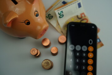 Several euro bills and several piles of coins of different currencies next to a piggy bank and a calculator