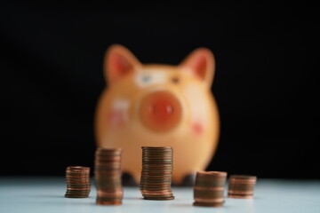 Several stacks of coins of different currencies in front of a piggy bank out of focus in the background as a savings concept