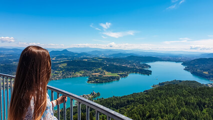 Tourist woman with panoramic view of idyllic lake Woerthersee in summer seen from observation tower Pyramidenkogel, Carinthia, Austria. Lakeside village surrounded by hills and mountains Austrian Alps