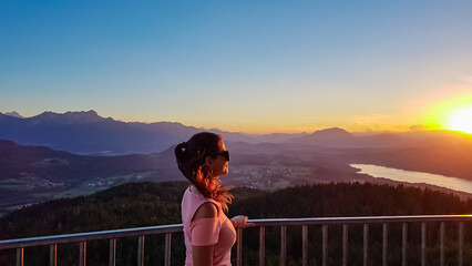 Fototapeta premium Happy woman with panoramic view of lake Woerthersee in summer during sunset seen from observation tower Pyramidenkogel, Carinthia, Austria. Romantic atmosphere surrounded by mountains of Austrian Alps