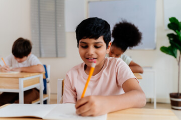 Happy Indian boy in a classroom setting, smiling brightly while writing. Young student with afro hair, sitting at a desk with a pencil and notebook