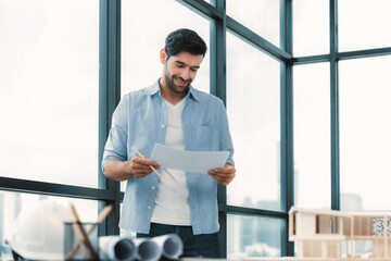 Expert civil engineer holding blueprint while comparing with house model on table with architectural equipment, document,blueprint house model scatter around. Real estate design concept. Tracery.