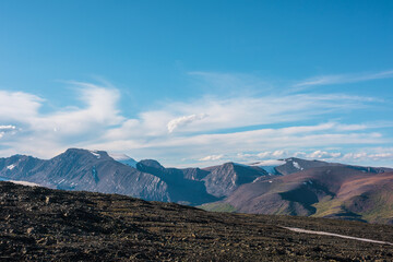 Top view from sunlit stony plain to large multicolor rocky mountains far away in bright sun under clouds in blue sky. Big colorful mountain range of many vivid colors in sunny day. Layered landscape.