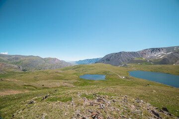 Two lakes, sunlit green meadow, alpine valley and mountain range. Blue sky gradient reflected in water. Top view from rocks to dirt road between two beautiful lakes in high mountains in sunny day.