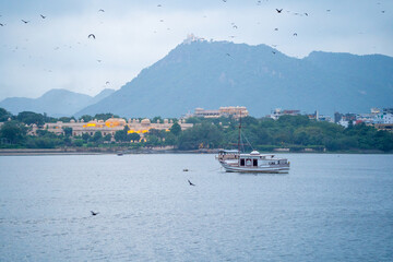 Dusk shot of lake pichola with boats moving and Leela palace oberoi in distance with Sajjan Garh monsoon palace in distance on hilltop showing aravalli hills the beauty of Udaipur