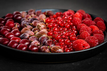 A mixture of red summer berries on black plate, angle view. Tasty fresh ripe red currant, raspberry, gooseberry, cherry berries. Healthy food texture on dark background.