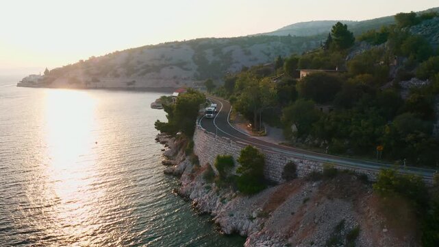 Minivan follows tourist minibus along highway at twilight. Route winds along slope near coastline framed by mountains and forests aerial view