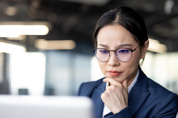 A focused asian businesswoman working on her laptop in a modern office setting. She is wearing a...