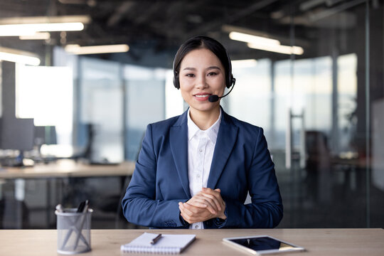 Professional businesswoman in formal attire communicating through a video call in a modern office environment. She is looking at the camera and wearing a headset, conveying professionalism