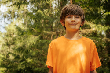 A young boy with brown hair and an orange shirt gazes intently in a forest setting. The background features lush greenery, capturing a moment of wonder and curiosity. © Emvats