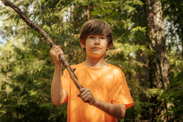 A young boy wearing an orange shirt holds a stick while standing in a forest. The background is...