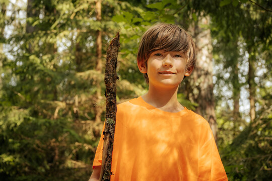 A young boy wearing an orange shirt holds a stick while standing in a forest. The background is filled with lush greenery, highlighting a moment of exploration and adventure in nature. - Powered by Adobe
