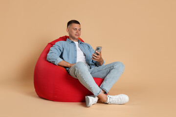 Handsome man with smartphone on red bean bag chair against beige background