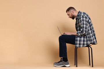 Man with poor posture sitting on chair and using laptop against pale orange background, space for text