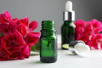 Bottles of geranium essential oil, pipette and beautiful flowers on white wooden table, closeup