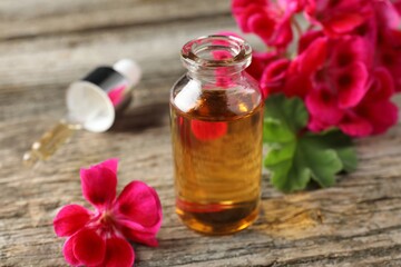 Bottle of geranium essential oil, pipette and beautiful flowers on wooden table, closeup