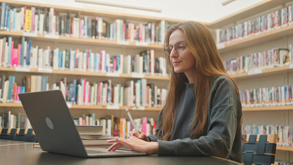 A smart college student uses a laptop to learn at the public library. Beautiful young Caucasian woman with glasses online studying, prepares for exams, takes notes with a pencil and writes essays 