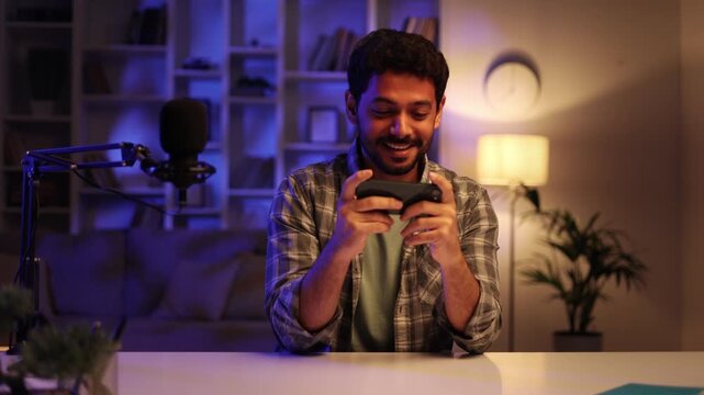 Young confident Arab man playing online video game on his phone, streaming live at home at night, talking into microphone. Gamer lifestyle, E-Sport online gaming technology concept.