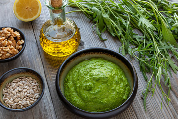 Wild arugula pesto in a bowl. On the side are the main ingredients: wild arugula leaves, walnuts, sunflower seeds, lemon, and olive oil.