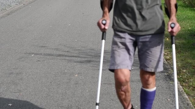 man with broken left leg in modern blue fiberglass plaster cast with walking shoes, walking with two legs and leaning on crutches, taking the load off the injured leg, rehabilitation concept,