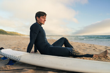 A surfer in a wetsuit sits on the sandy beach beside his surfboard, enjoying the serene ocean view...
