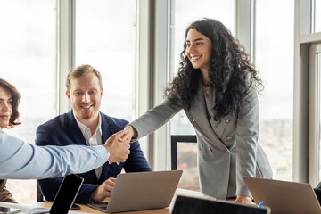 A businesswoman in a grey blazer leans across a conference table to shake the hand of a male...