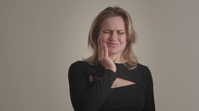 A young american or european unhealthy blonde woman suffers horrible toothache or gumboil, supports and massages cheek, tilts head aside and frowns face in pain. Teeth decay, inflammation or dental