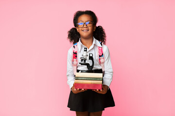 Unhappy Elementary School Girl Holding Heavy Books And Microscope Standing Over Yellow Studio Background. Too Much Quarantine Homework.