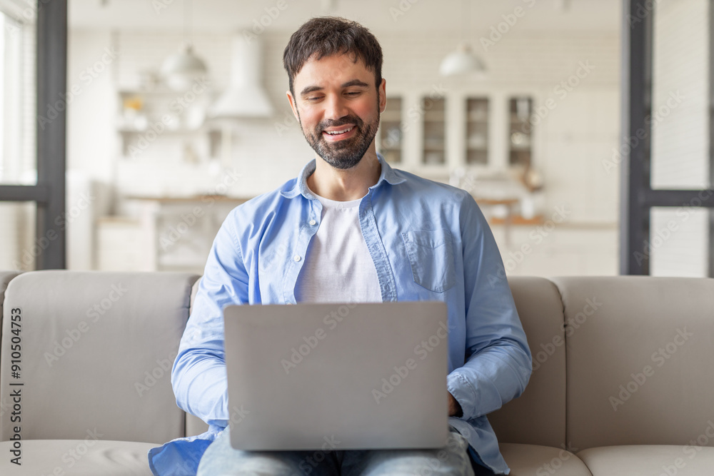Poster a man is sitting on a light grey couch in a modern living room, working on his laptop. he is smiling