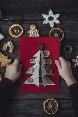 Idea for easy Christmas do it yourself crafts for kids to make. Child's hands with handmade postcards with fir tree and toy from recycled cardboard paper, thread, buttons. Wooden background, flat lay