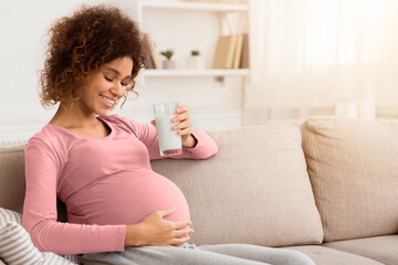 Healthy pregnancy. African expectant woman holding glass of fresh milk, sitting on sofa, empty space